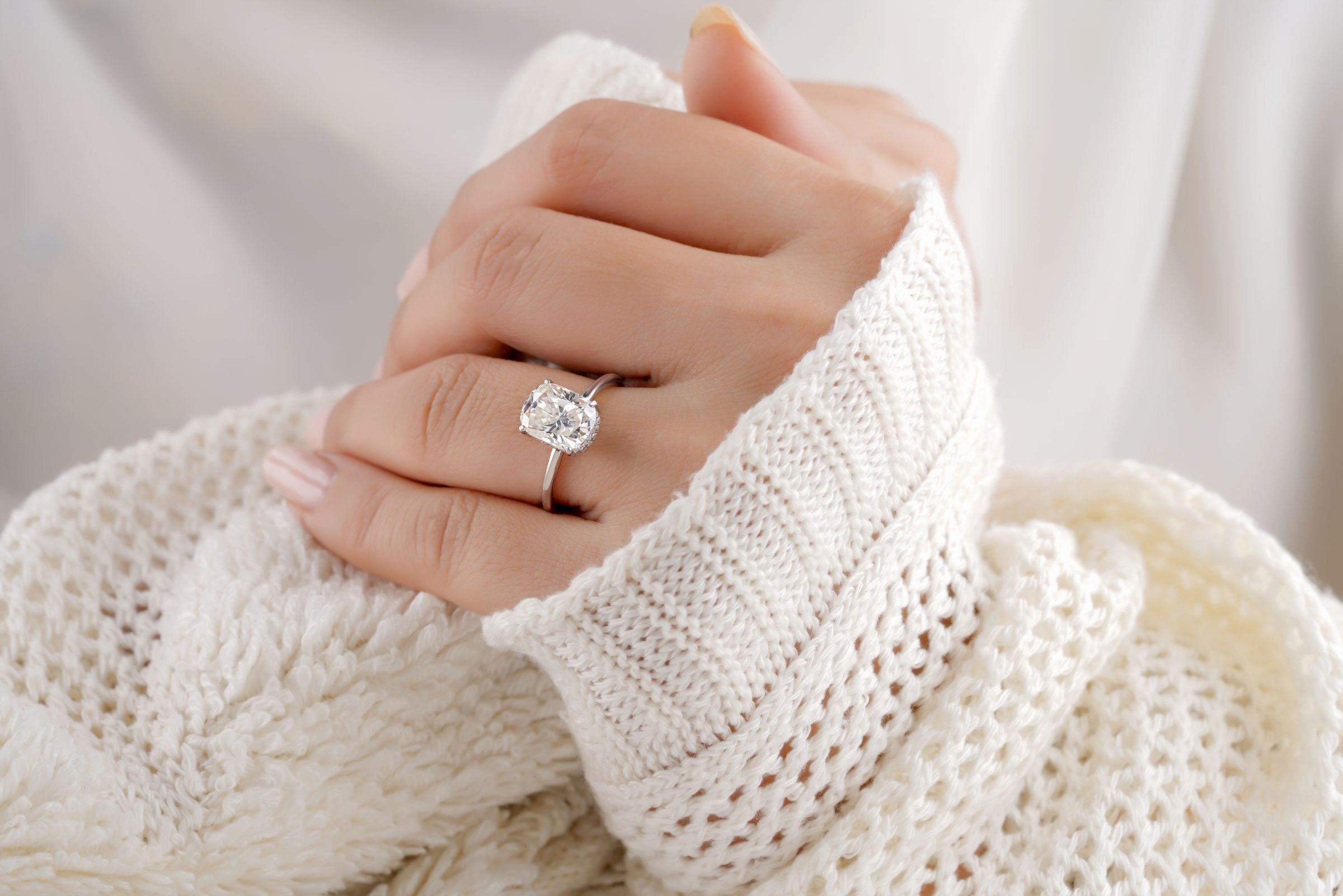 A hand wearing a cushion-cut diamond solitaire ring, wrapped in a cream knit sleeve